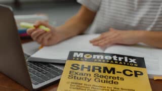 Exam prep book on table in front of person studying with laptop