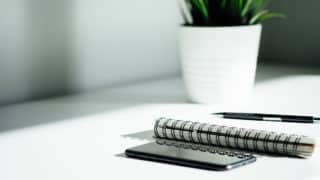 Smartphone, notebook, and pen on white table in front of potted plant